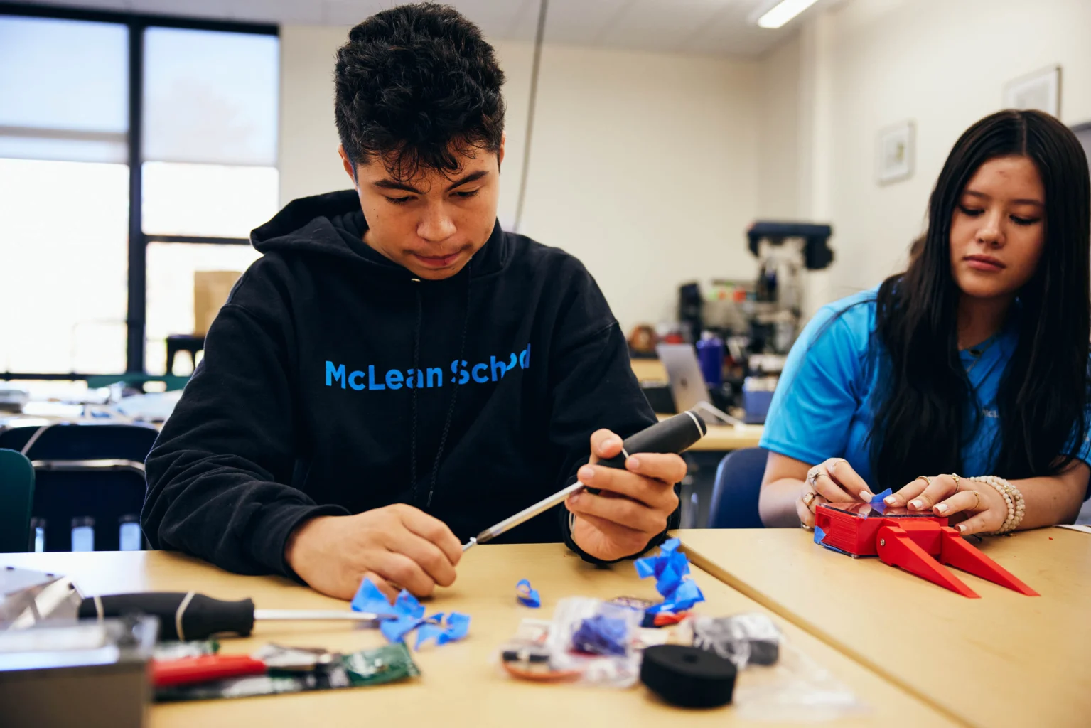 Two students sit at a classroom table working on hands-on projects with tools and electronic components in front of them.