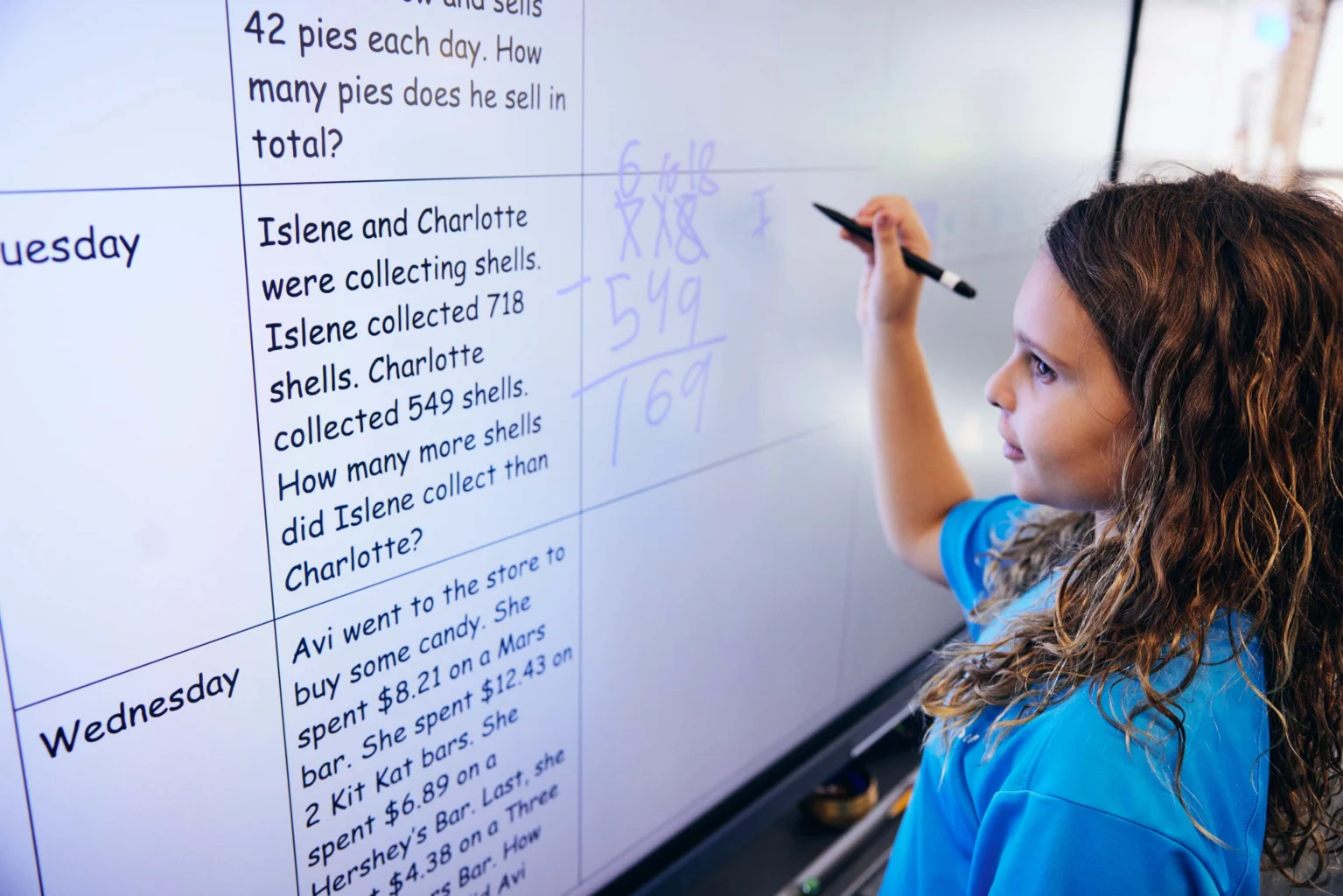 A girl writes math calculations on a large interactive whiteboard, solving a word problem about collecting shells.