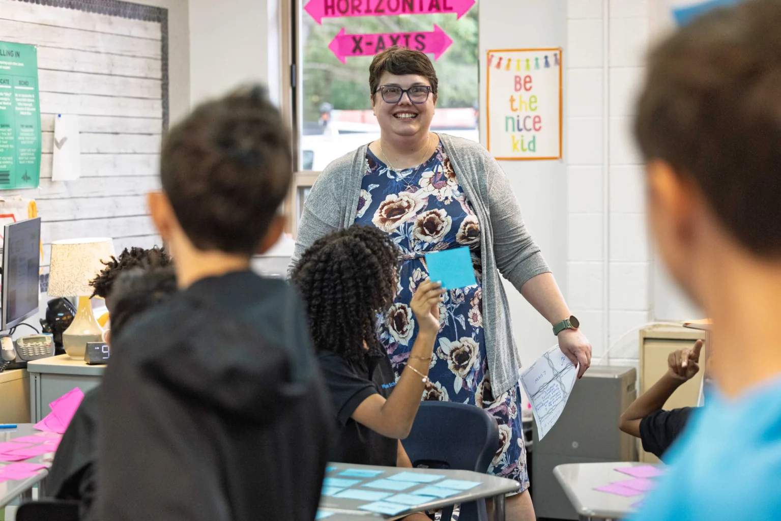A teacher stands and smiles in a classroom as students participate, one holding up a blue sticky note. Classroom signs and posters are visible in the background.