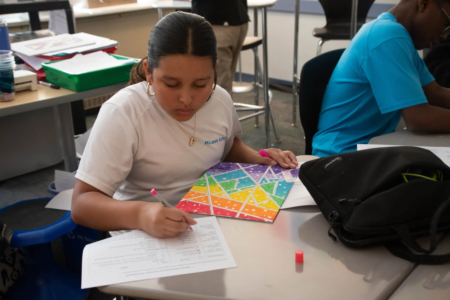 Student in a white shirt completes a worksheet at a desk, holding a pink pen and a colorful geometric board, with school supplies nearby.