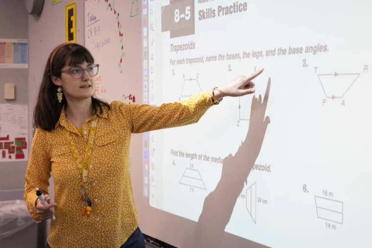 A woman in a yellow shirt points at a projected math lesson about trapezoids on a whiteboard in a classroom.