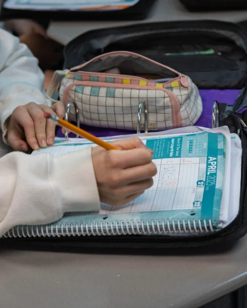 A person writes in a planner with a pencil at a desk, next to an open binder and a pencil case.