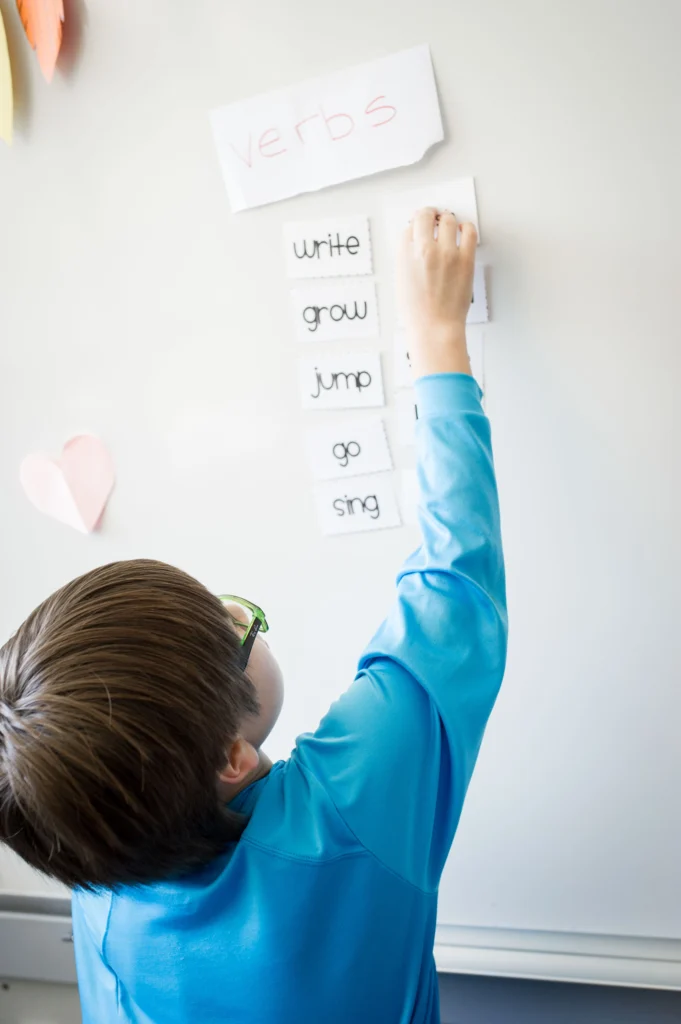 A child in a blue shirt reaches up to attach a flashcard to a whiteboard labeled "verbs," with other verb cards like "write," "grow," "jump," "go," and "sing" displayed.