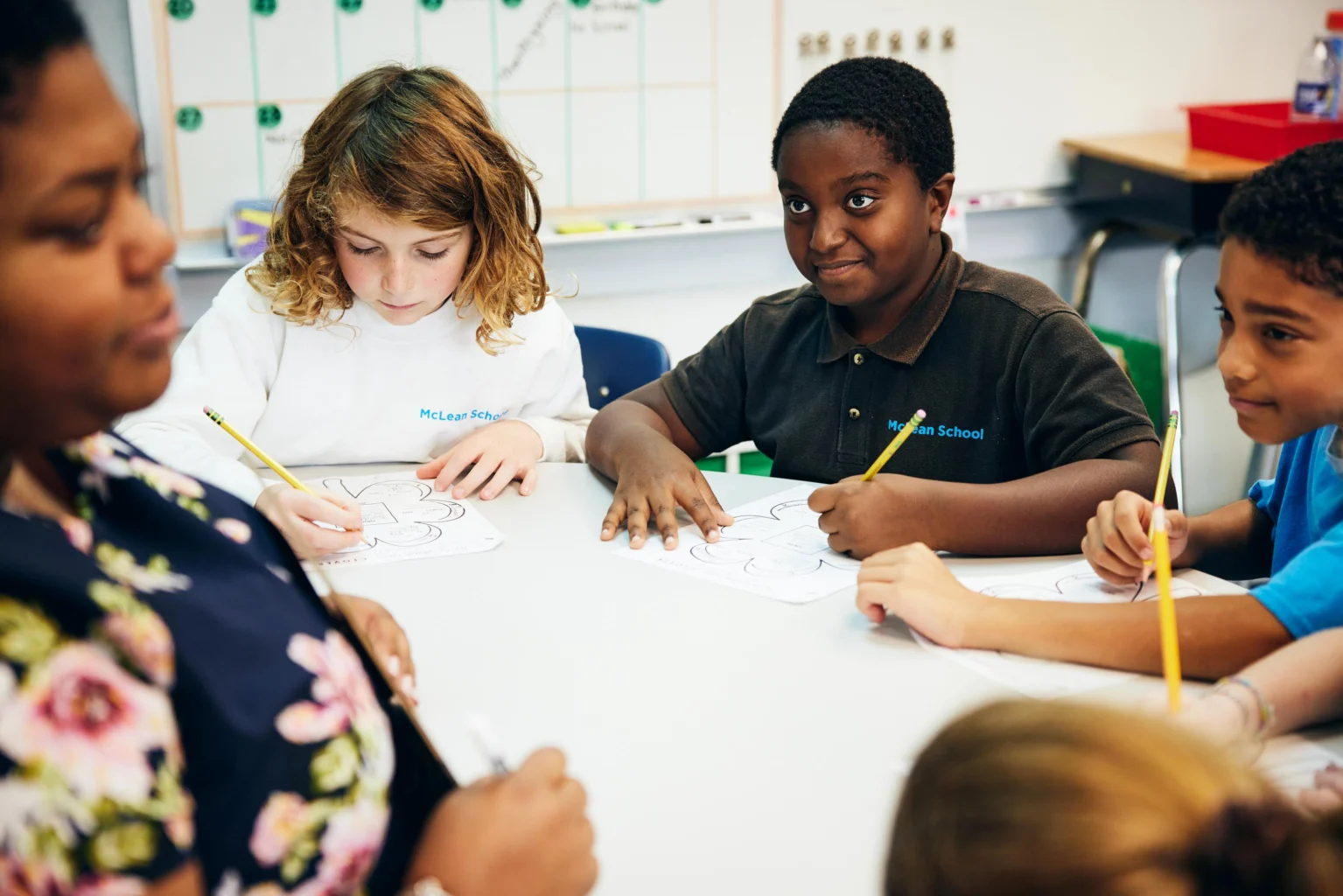 A teacher sits with a group of elementary school students at a table as they work on worksheets and write with pencils in a classroom.