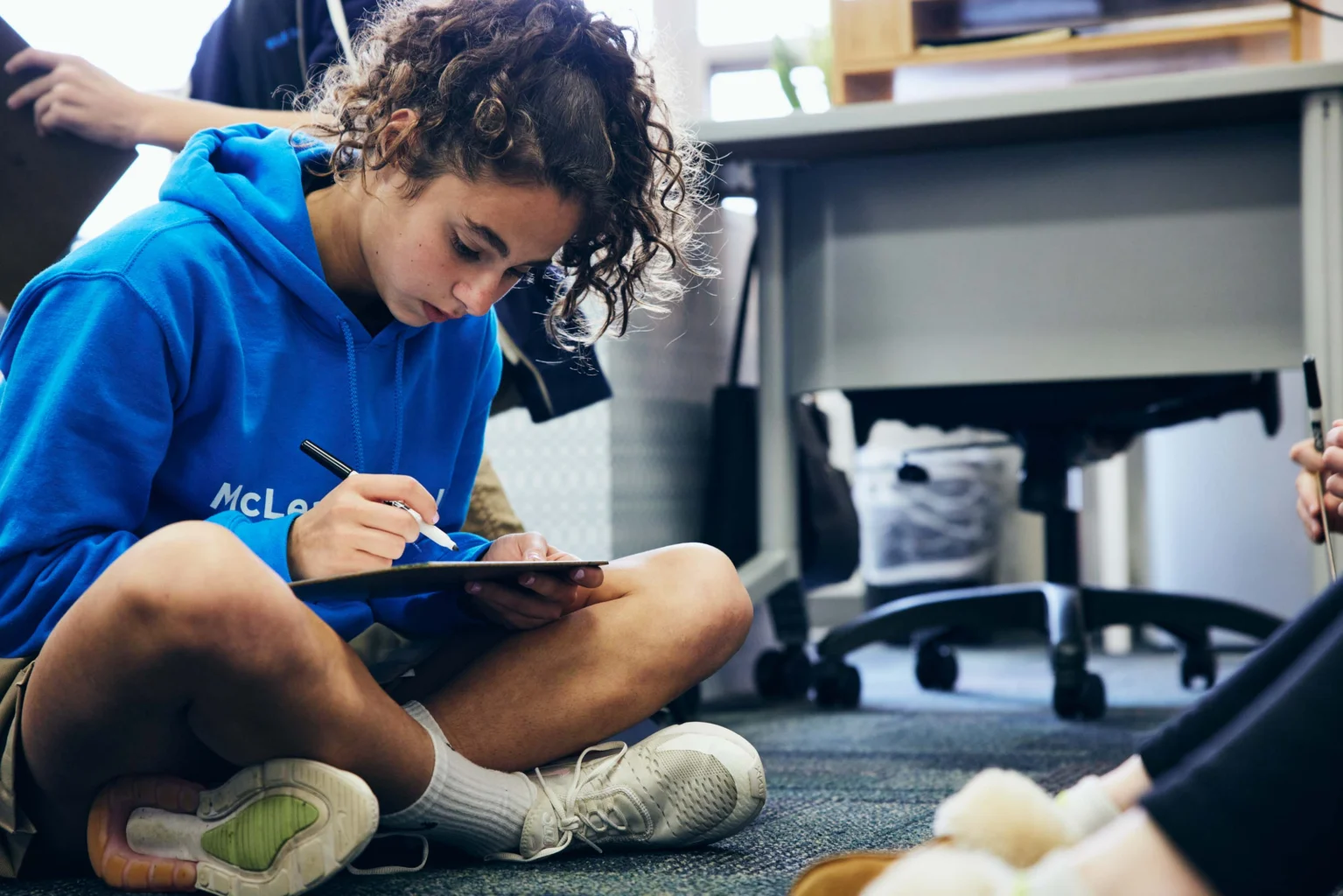 A young person in a blue hoodie sits cross-legged on the floor, writing on a clipboard, with desks and other people nearby.