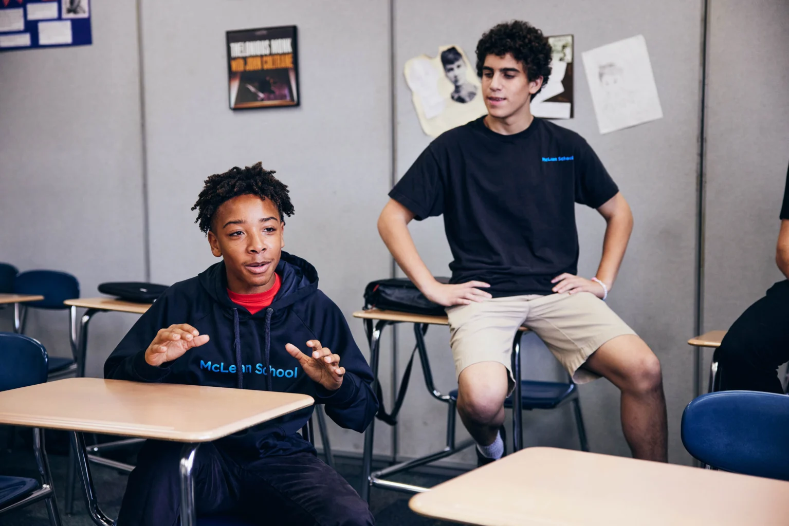 Two male students in a classroom, one seated and speaking while gesturing with his hands, and the other sitting casually on a desk, both wearing dark shirts.