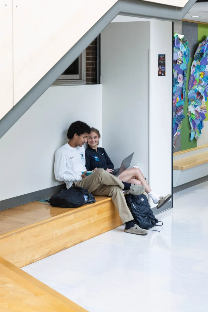 Two students sit on a wooden bench under a staircase; one holds a phone while the other uses a laptop. Backpacks are on the floor beside them. Colorful artwork hangs on the wall nearby.