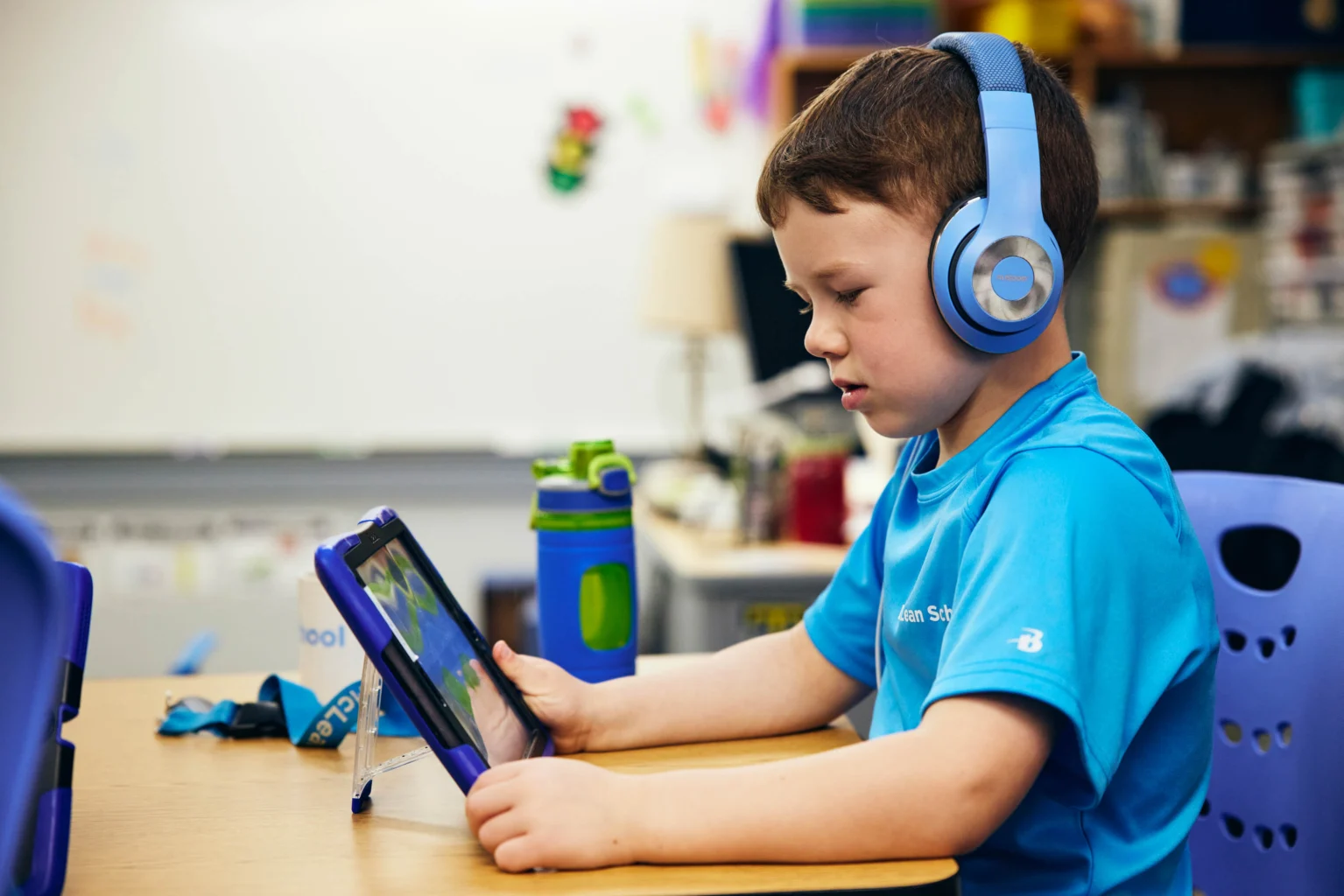 A young boy wearing headphones uses a tablet at a classroom desk, with a water bottle and lanyard nearby.