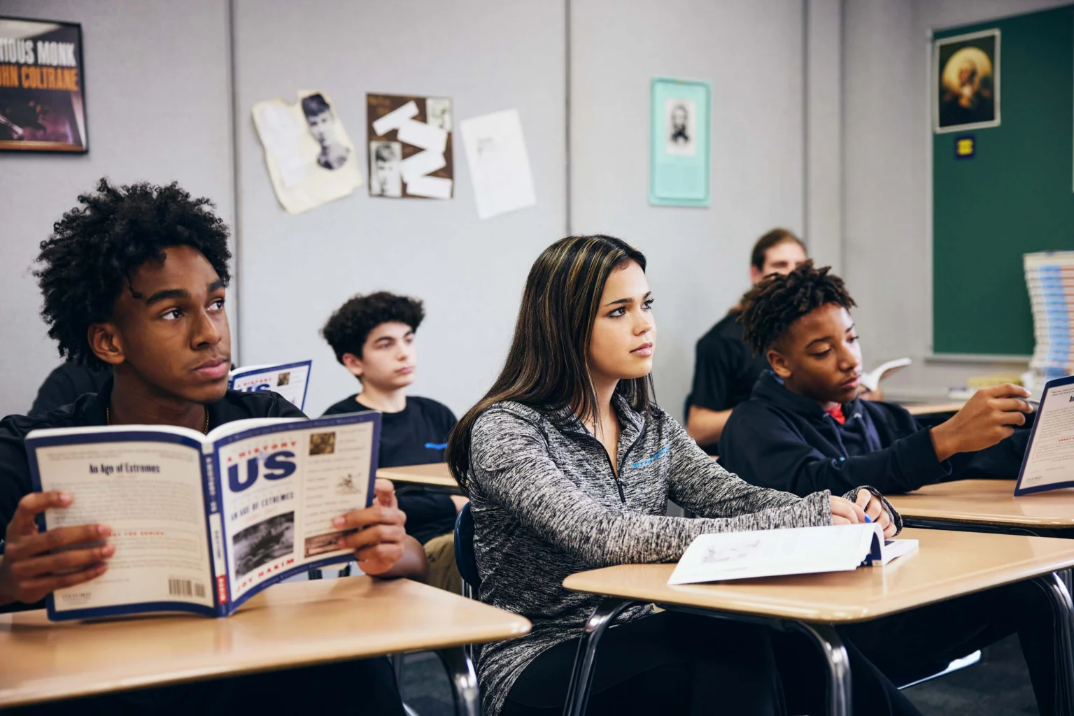 A group of students sit at desks in a classroom, reading textbooks and taking notes during a lesson.
