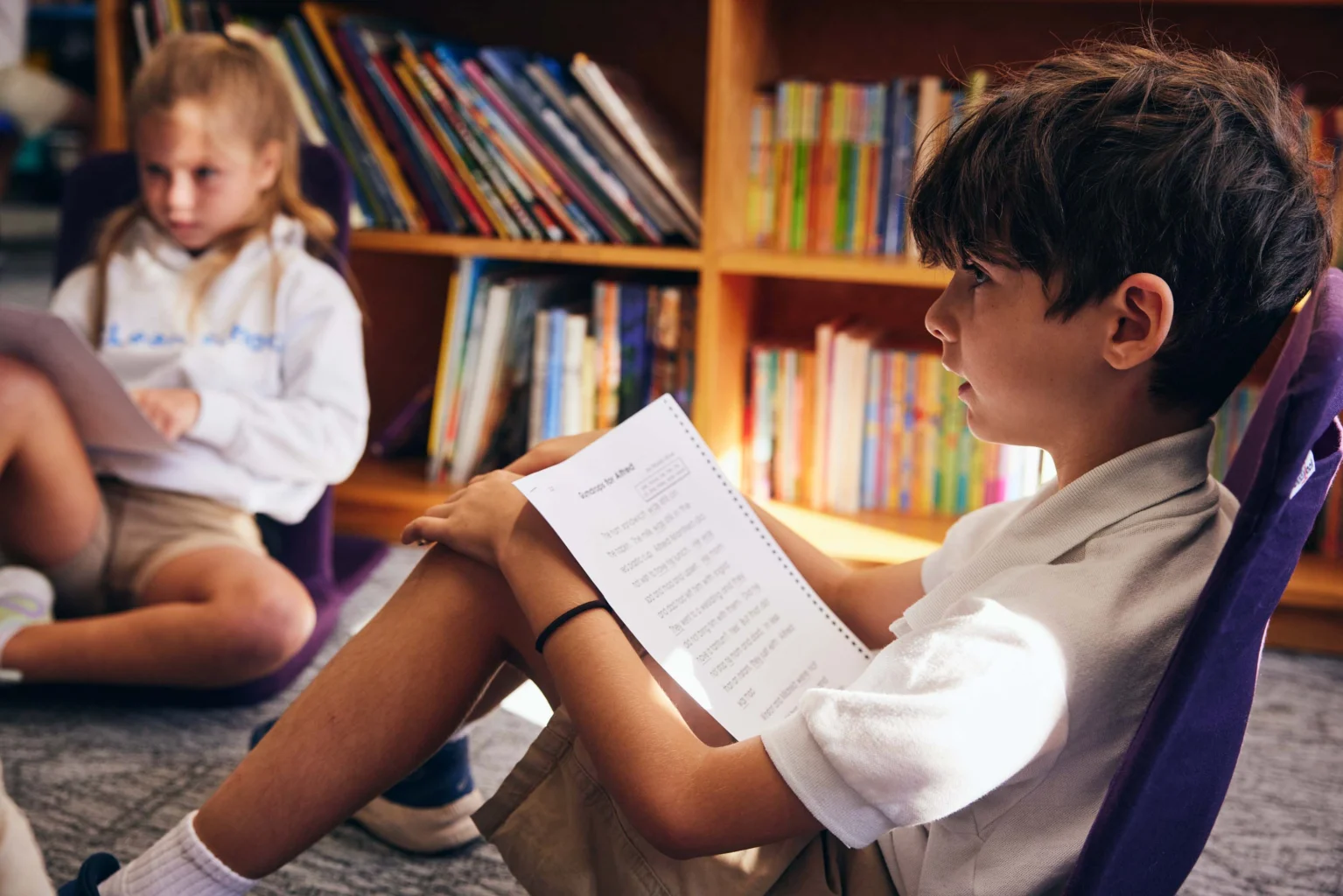 Two children sit on the floor in front of bookshelves; the boy in the foreground reads a spiral-bound book, while the girl in the background holds a notebook.