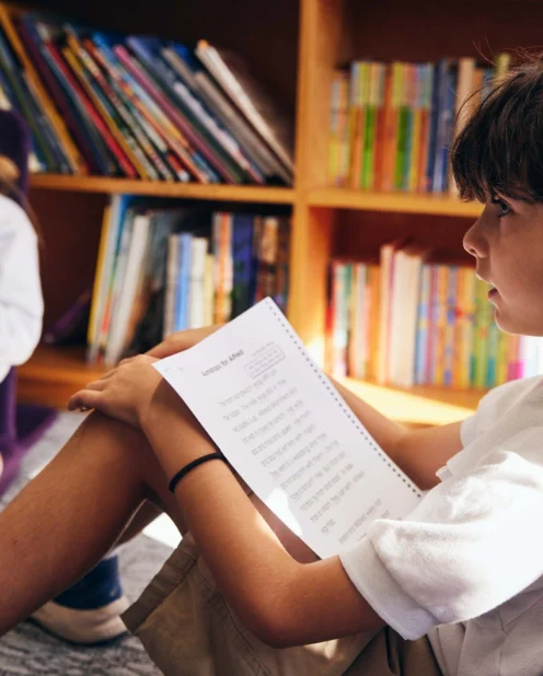 Two children sit on the floor in front of bookshelves; the boy in the foreground reads a spiral-bound book, while the girl in the background holds a notebook.