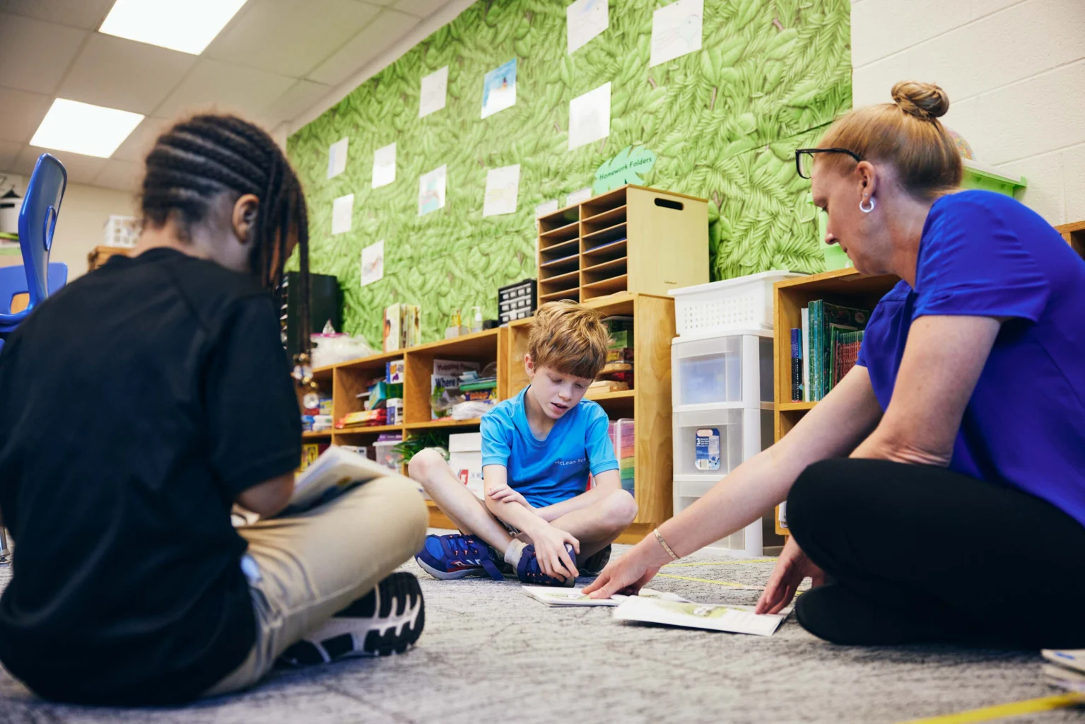 Three people sit on a classroom floor; a teacher points to a paper while two students look at materials, with shelves and green wall decor in the background.