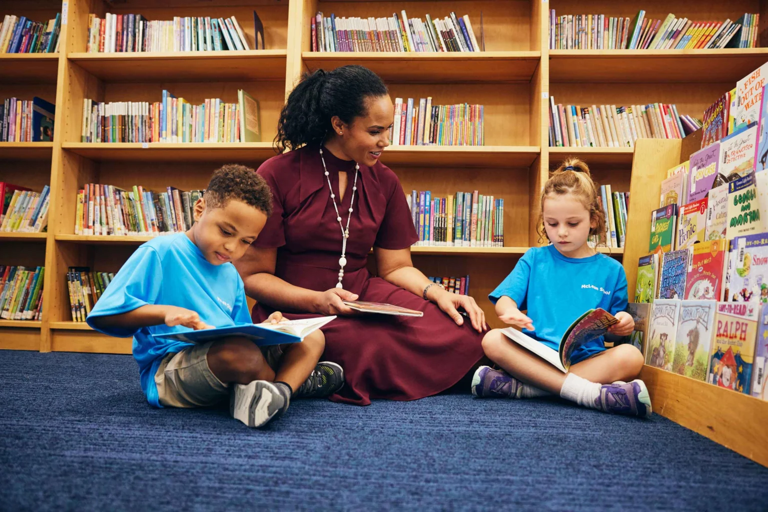 A woman sits on the floor in a library with two young children, all reading books, surrounded by bookshelves filled with colorful books.