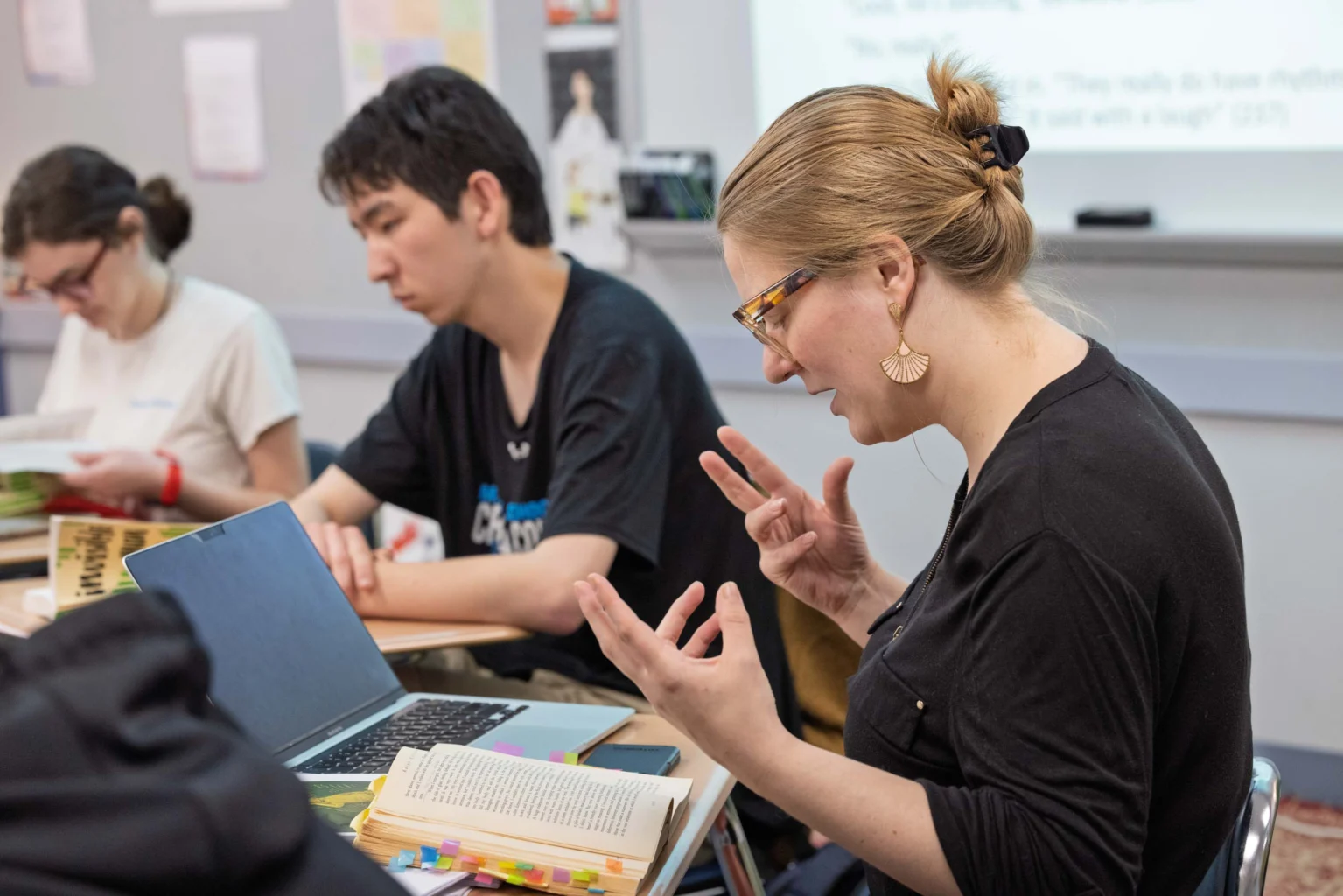 A teacher gestures while speaking to students in a classroom; an open laptop and annotated books are on the table.