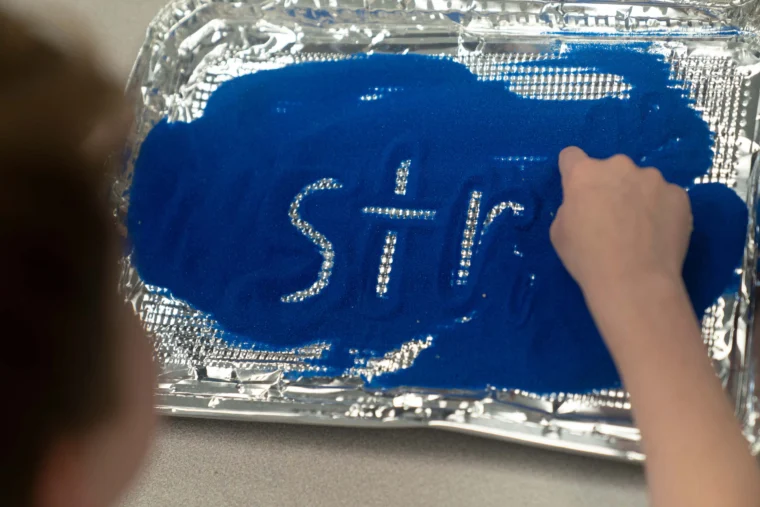 A child writes "Str" with their finger in blue sand spread on a foil-lined tray.