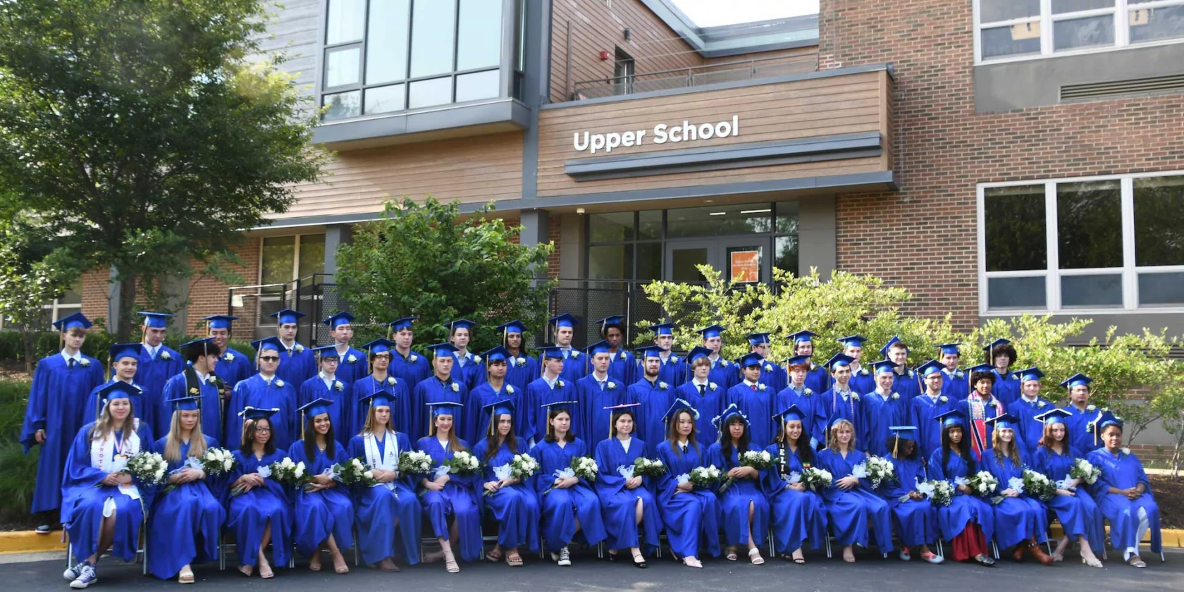 A group of students in blue graduation gowns and caps pose for a photo in front of a school building labeled "Upper School.