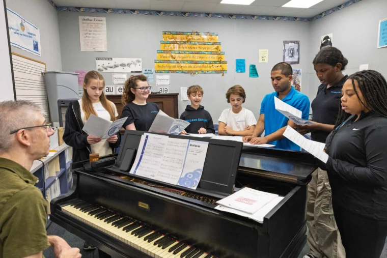 A group of students stand around a piano holding sheet music and singing while a teacher sits at the piano in a classroom.