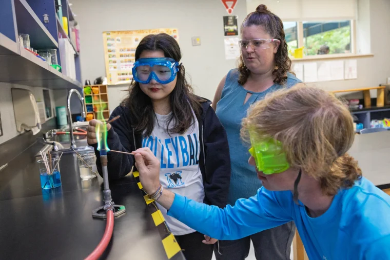 Three students in safety goggles conduct a science experiment with liquids in a classroom laboratory setting.