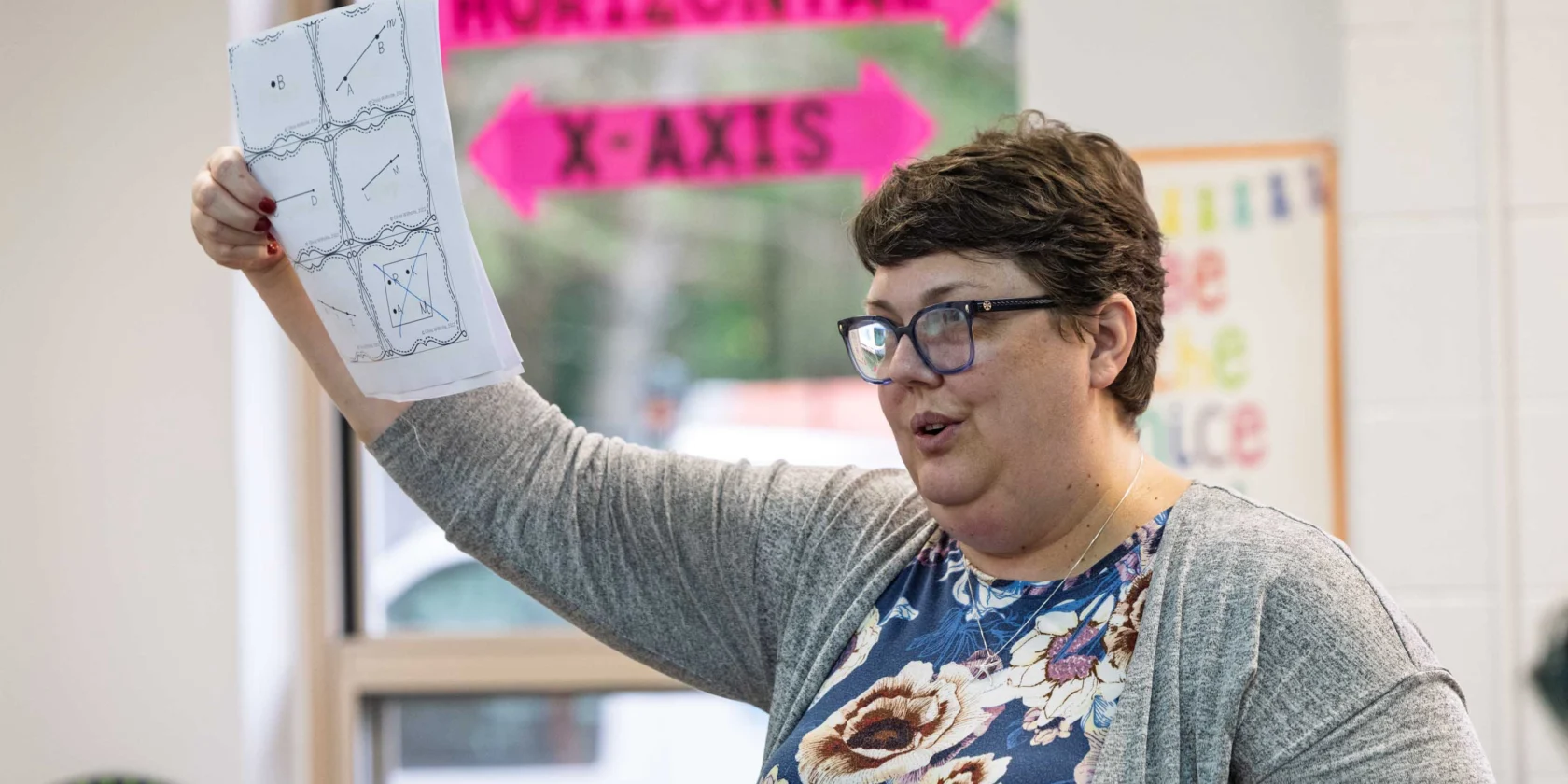 A woman wearing glasses holds up a paper with diagrams in a classroom. A bright pink sign reading "HORIZONTAL X-AXIS" is visible in the background.