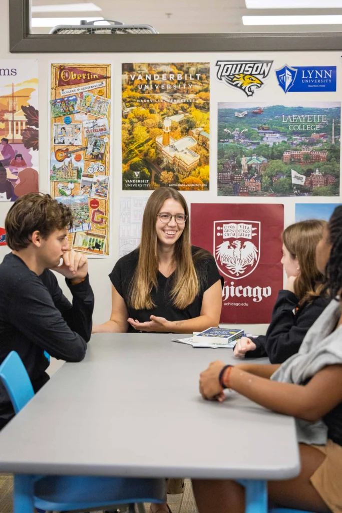 A woman sits at a table talking with three students in a classroom, with college posters displayed on the wall behind them.