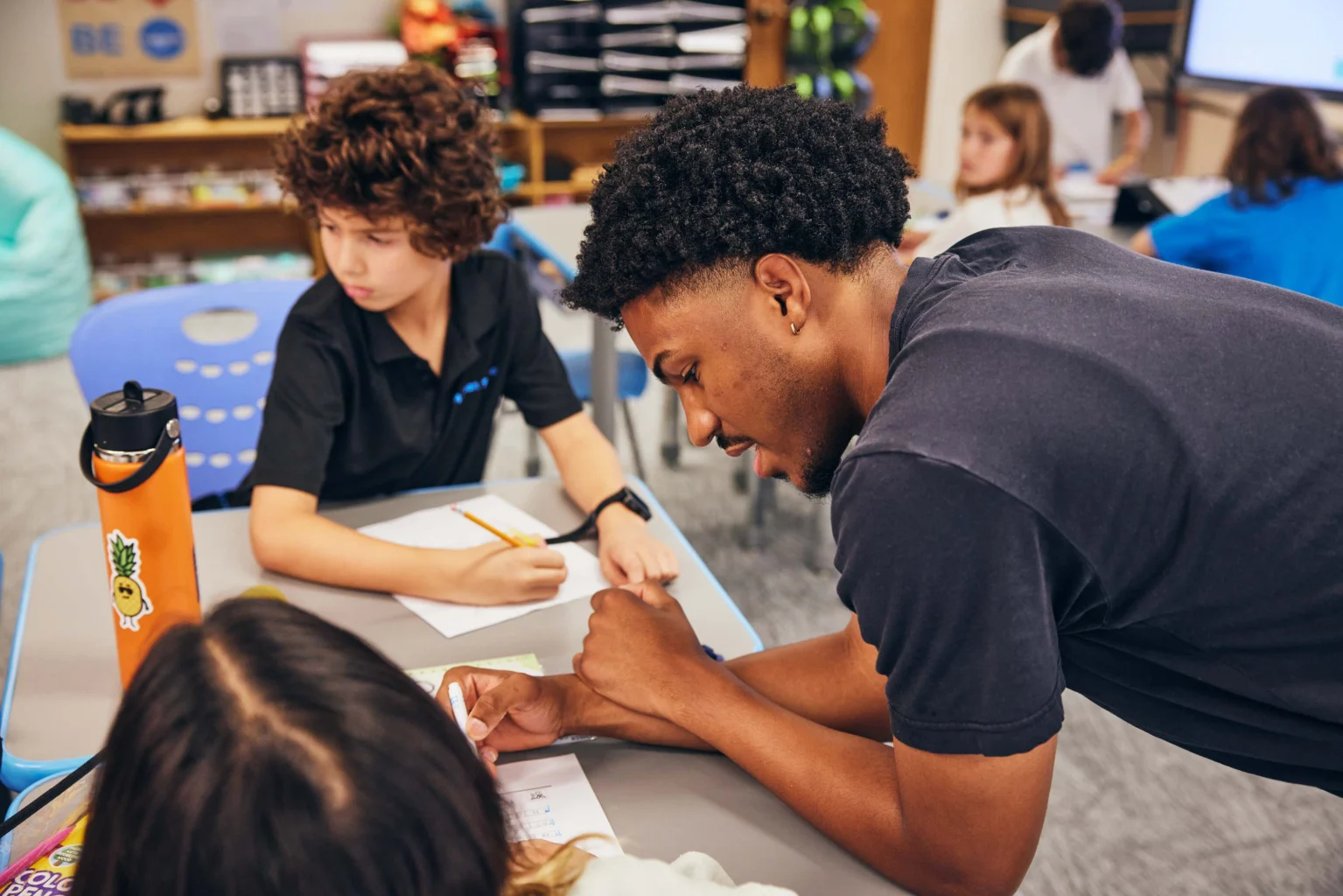 A teacher leans over a desk to assist a student with their work in a classroom while other students are seated and writing.