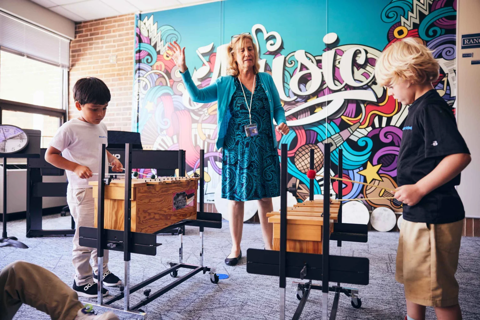 A teacher stands in front of a colorful music-themed wall, guiding two young boys playing xylophones in a classroom.