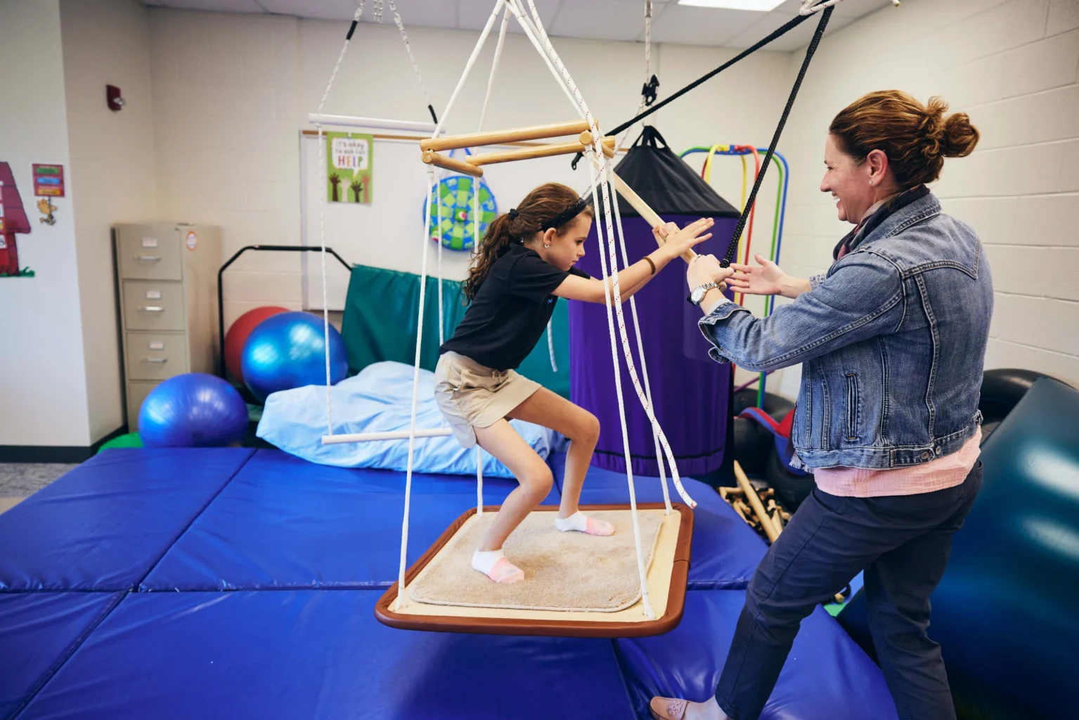A child stands on a suspended swing while an adult assists her in a sensory gym with mats, exercise balls, and equipment in the background.