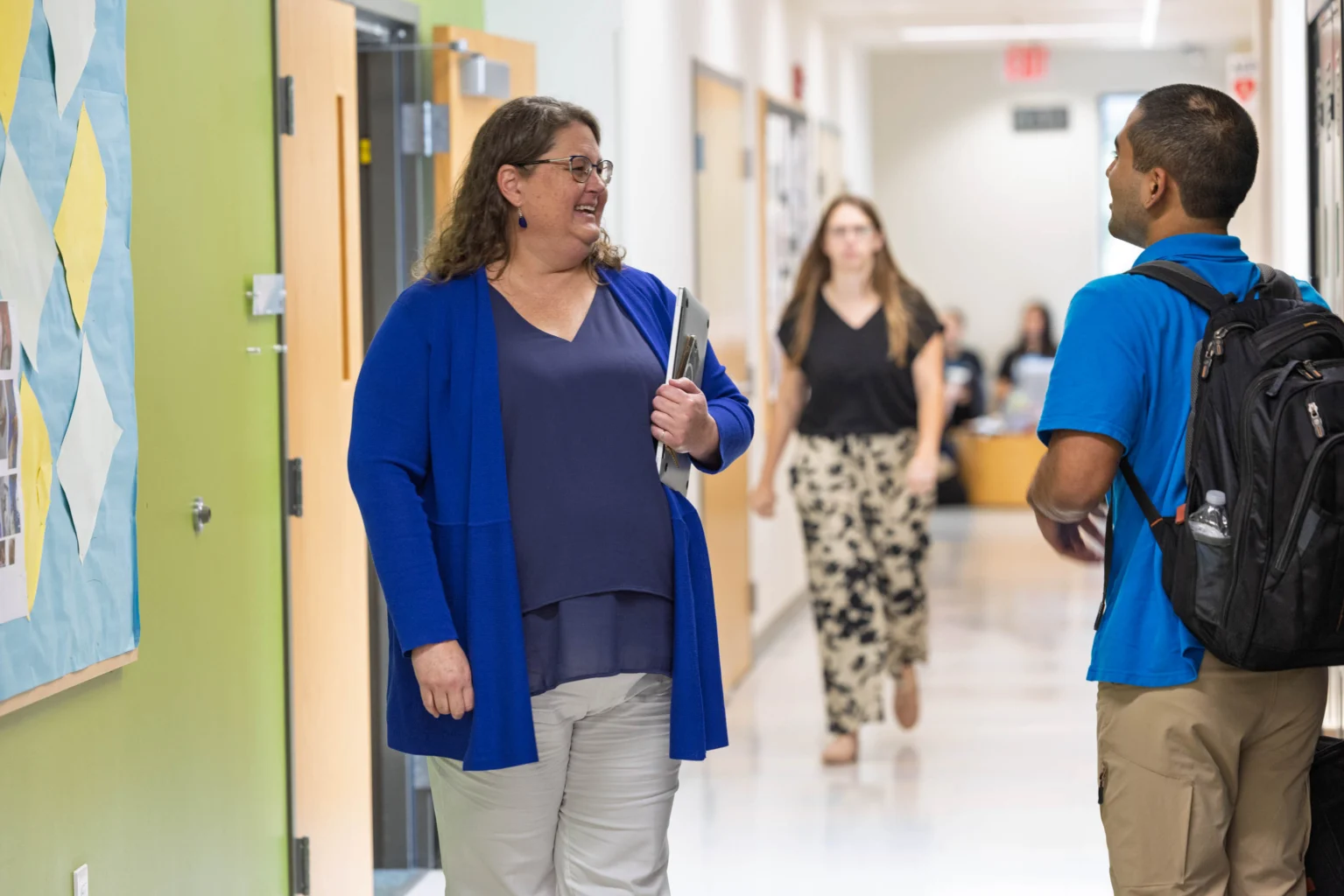 A woman holding a laptop and a man with a backpack talk in a school hallway while other people walk in the background.
