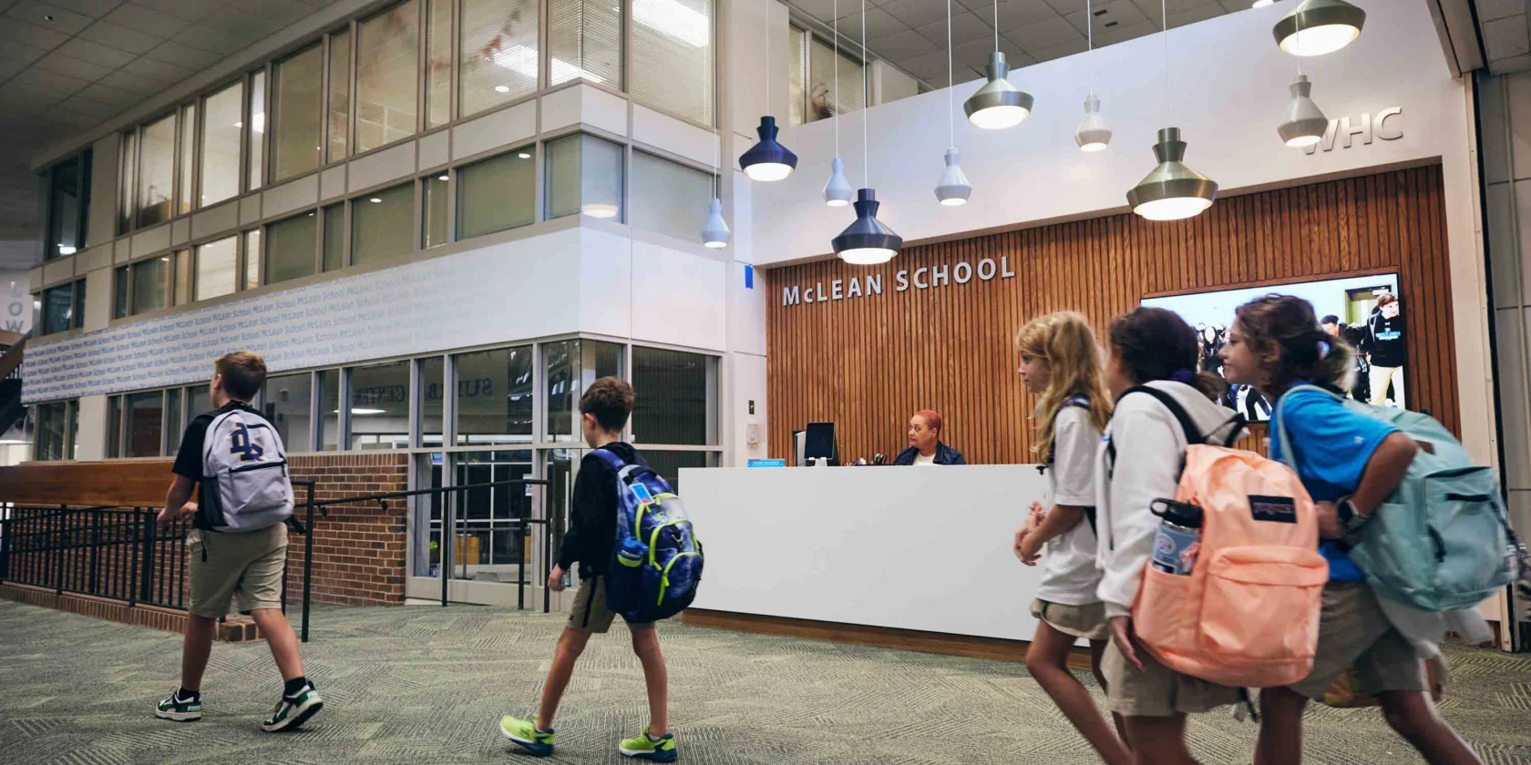 Four children with backpacks walk past the reception desk in the lobby of McLean School, where a staff member is seated behind the counter.