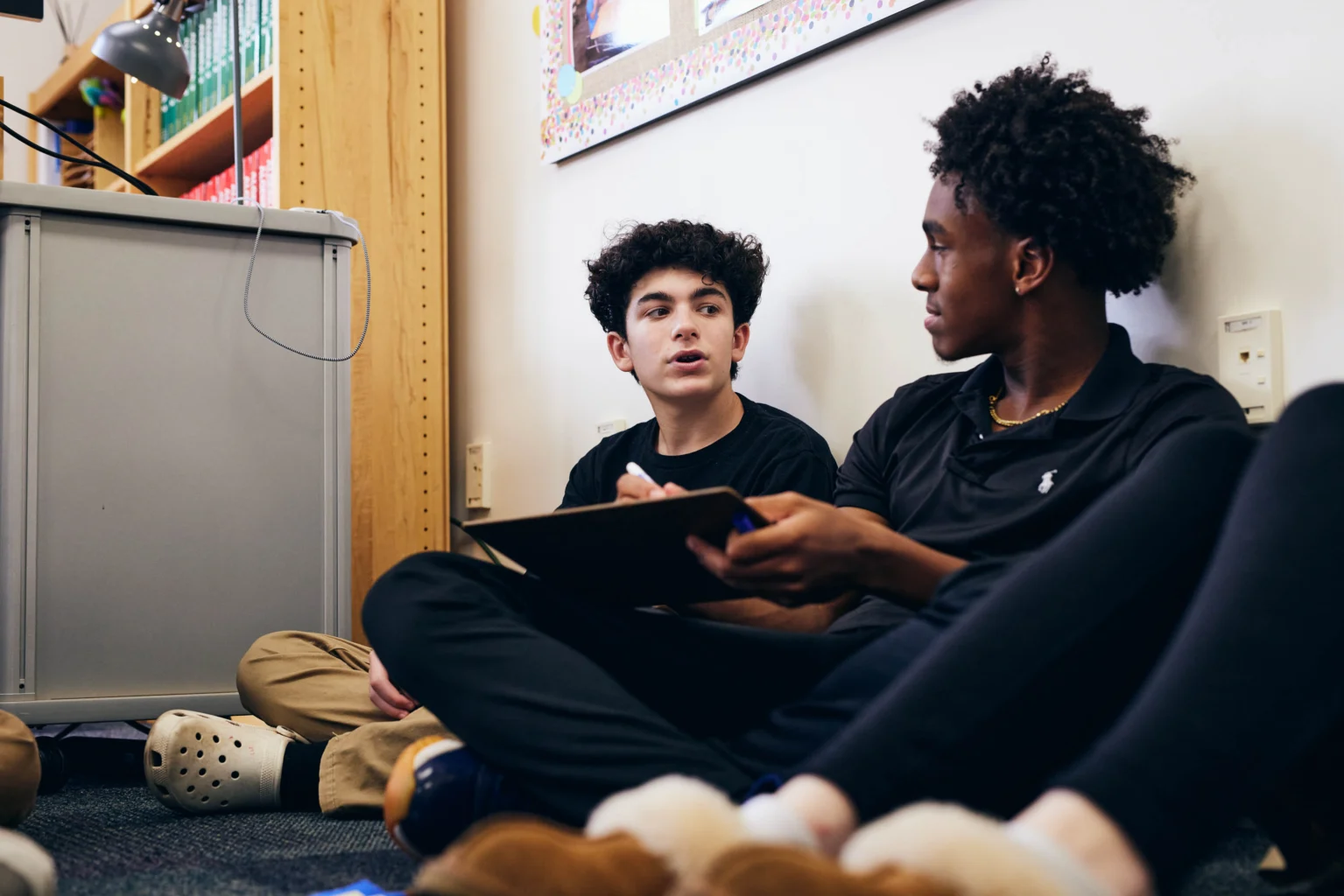 Two teenage boys sit on the floor indoors, facing each other and having a conversation; one holds a clipboard.
