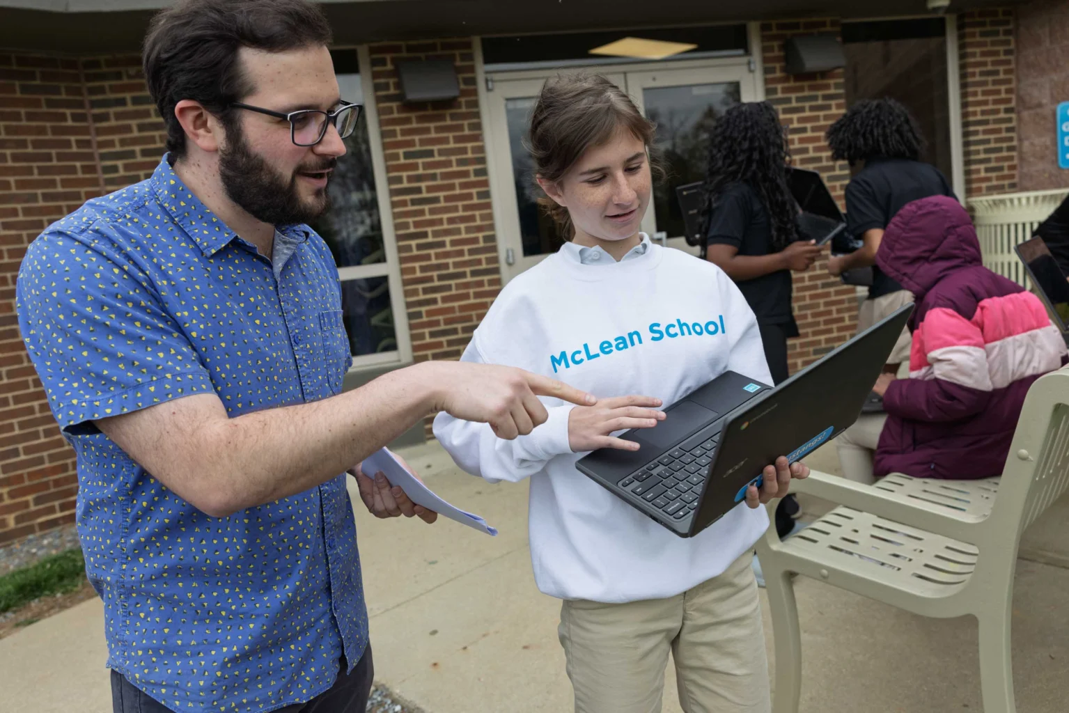 A bearded man points at a laptop screen held by a young woman in a "McLean School" sweatshirt outside a brick building. Other people are visible in the background.