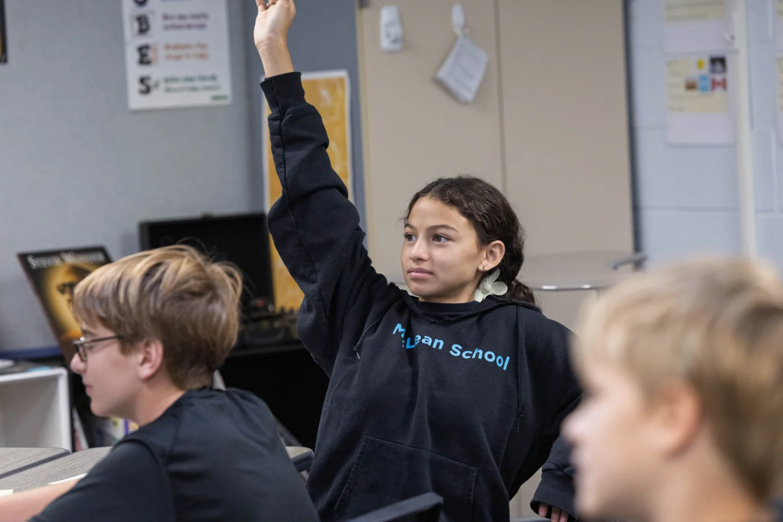 A student wearing a dark hoodie raises their hand in a classroom while other students sit nearby, facing forward.