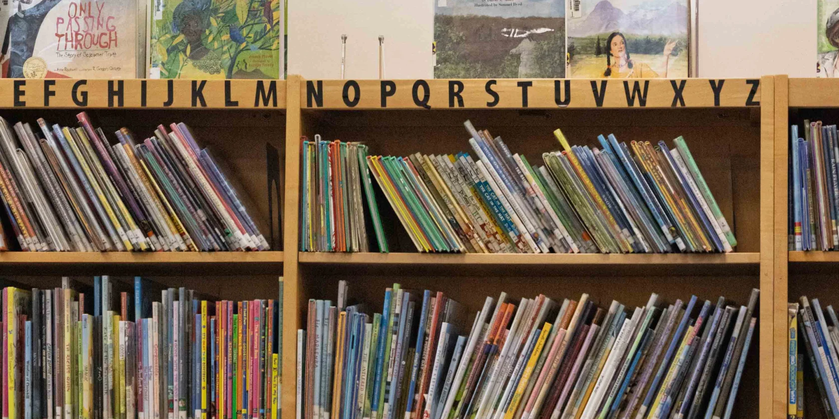Wooden bookshelves filled with books are organized alphabetically, with picture books displayed on the top shelf and letter labels visible above each section.