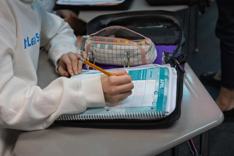 A person writes in a planner on a desk, with a pencil case and a binder nearby. The planner is open to a page for April.