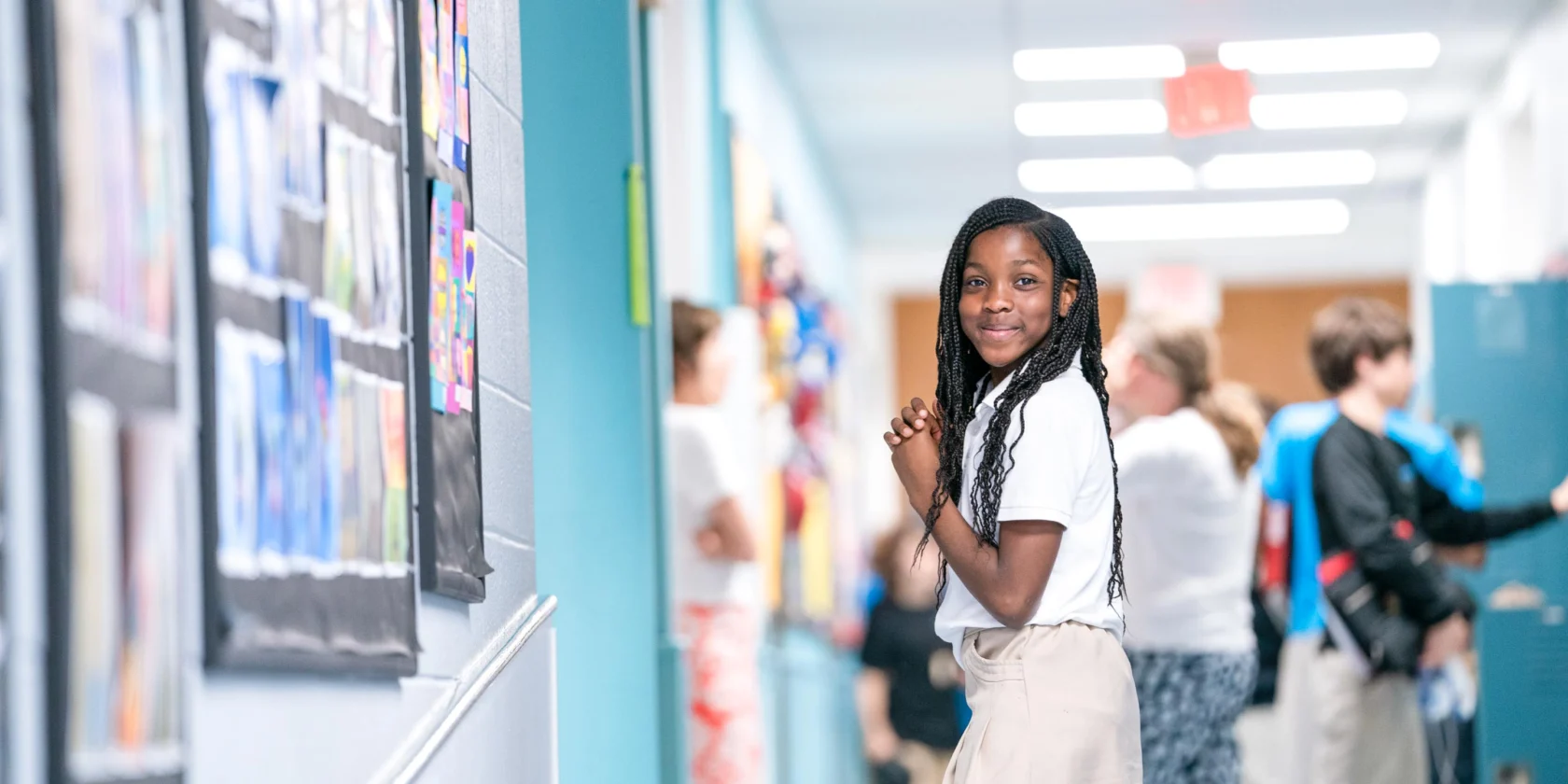 A student stands in a school hallway, smiling at the camera. Other students are nearby, some using lockers and some walking through the hallway.