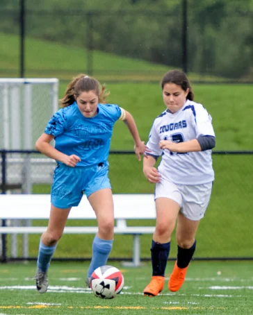Two female soccer players compete for the ball on a field, one wearing a blue uniform and the other in white, with a bench and green grass in the background.