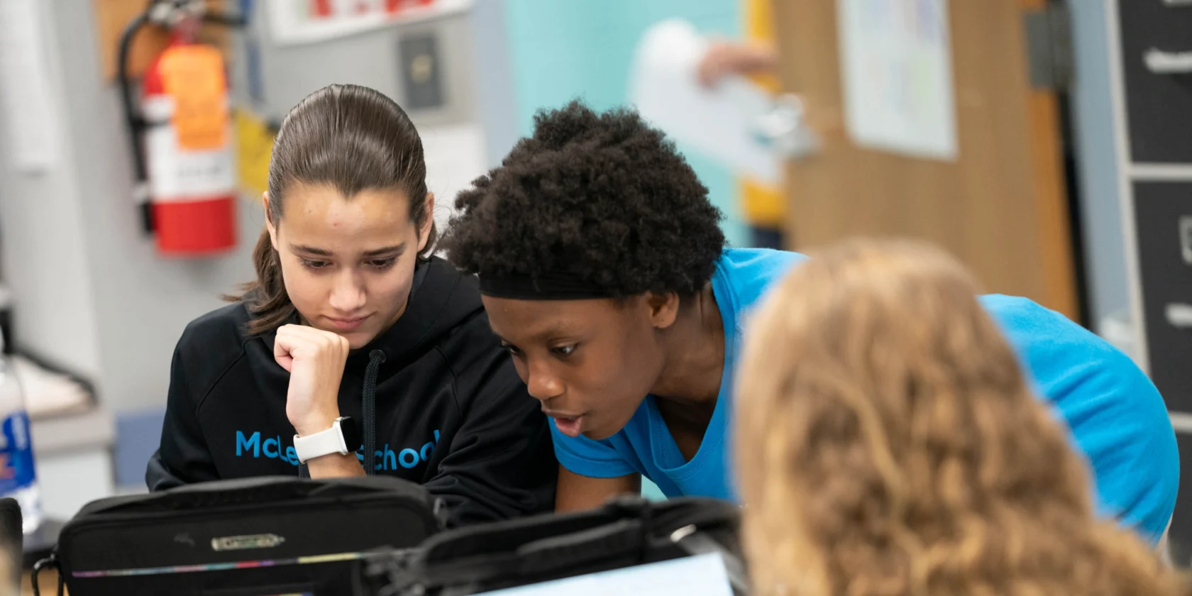 Three students work together at a table, looking at laptops in a classroom setting.