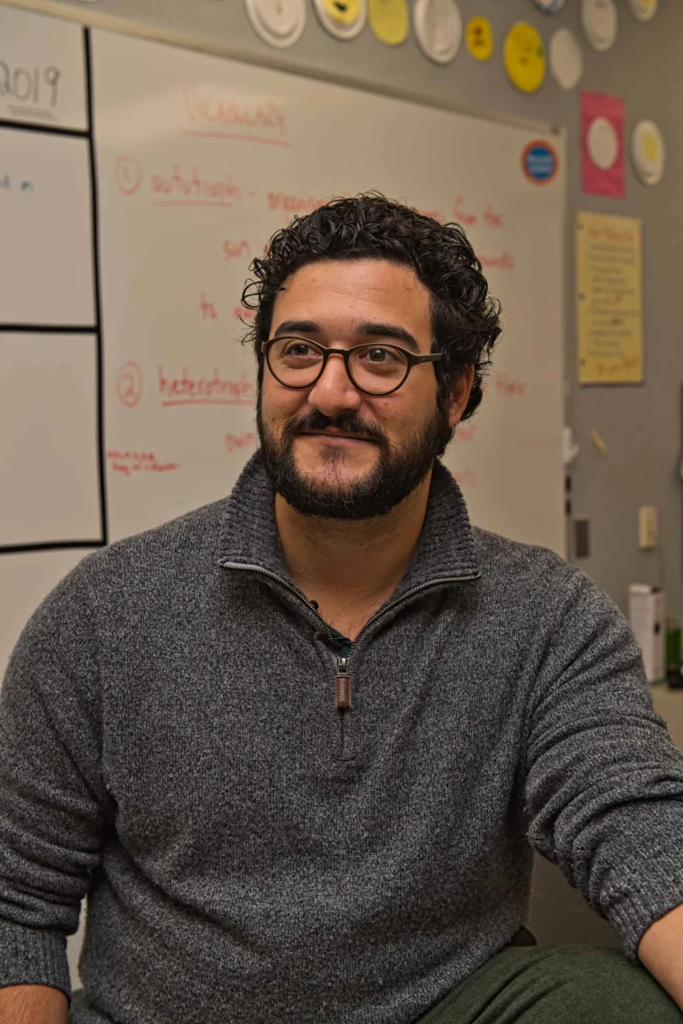 A man with dark curly hair, glasses, and a beard sits in a classroom, smiling slightly. A whiteboard with red writing and paper plates decorate the background wall.