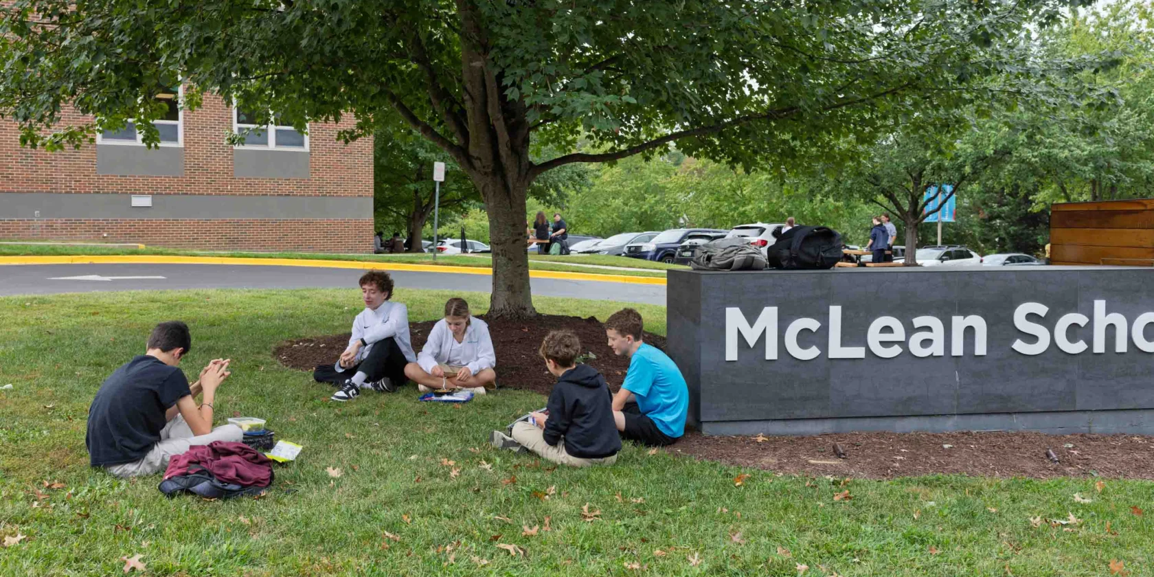 Five students sit on the grass near a tree, working together beside a large "McLean School" sign, with a brick building and parked cars in the background.