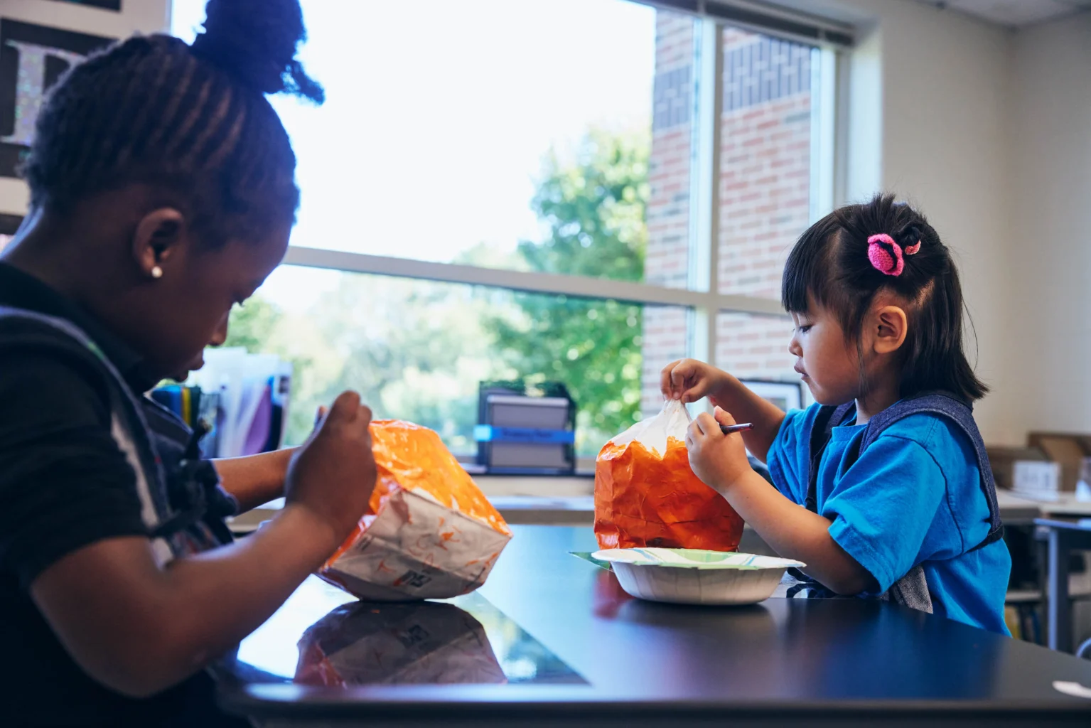 Two young children sit at a table in a classroom, eating food from bags and bowls with utensils.