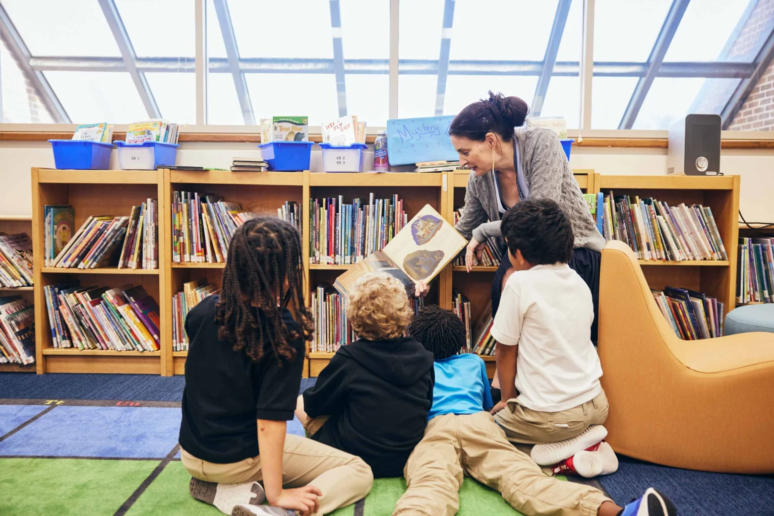 An adult reads a picture book to four children seated on the floor in front of bookshelves in a library.