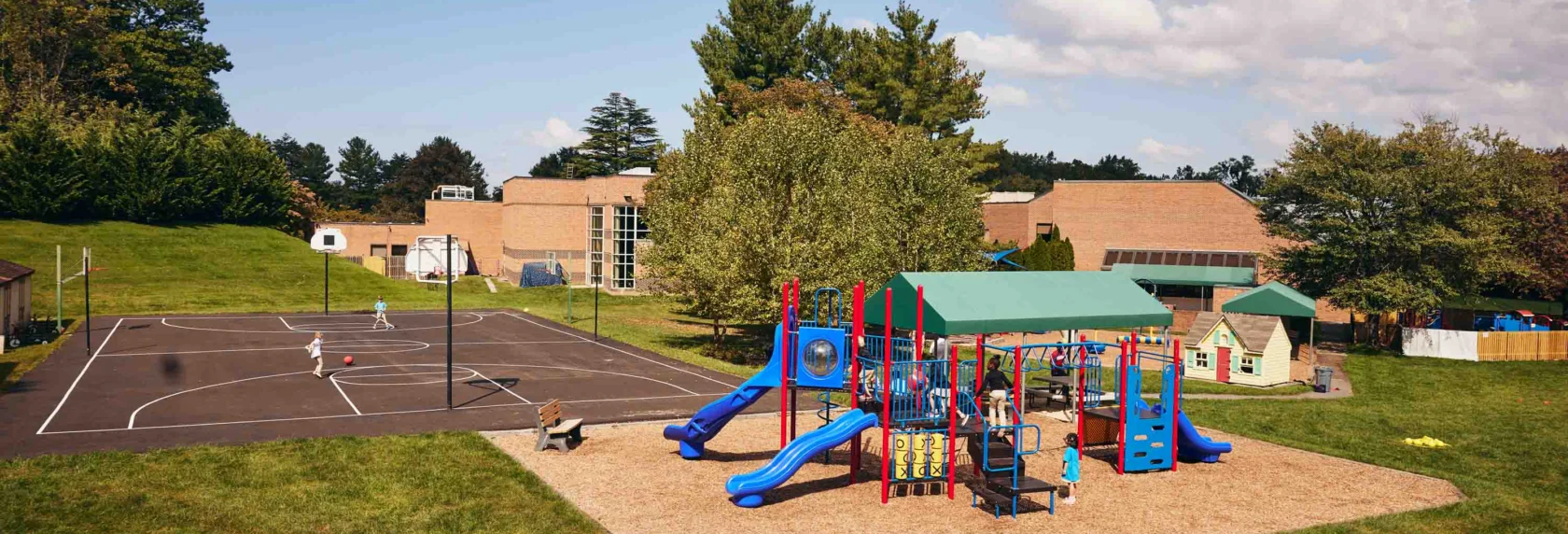 A playground with slides and climbing equipment sits next to a basketball court and a building, surrounded by grass and trees under a partly cloudy sky.