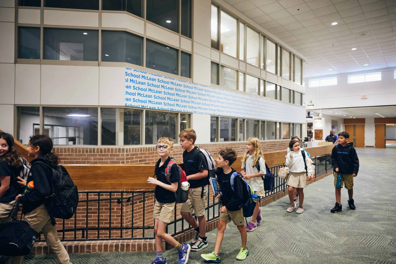 A group of elementary school children wearing backpacks walk in a line through a school hallway with large windows and brick accents.