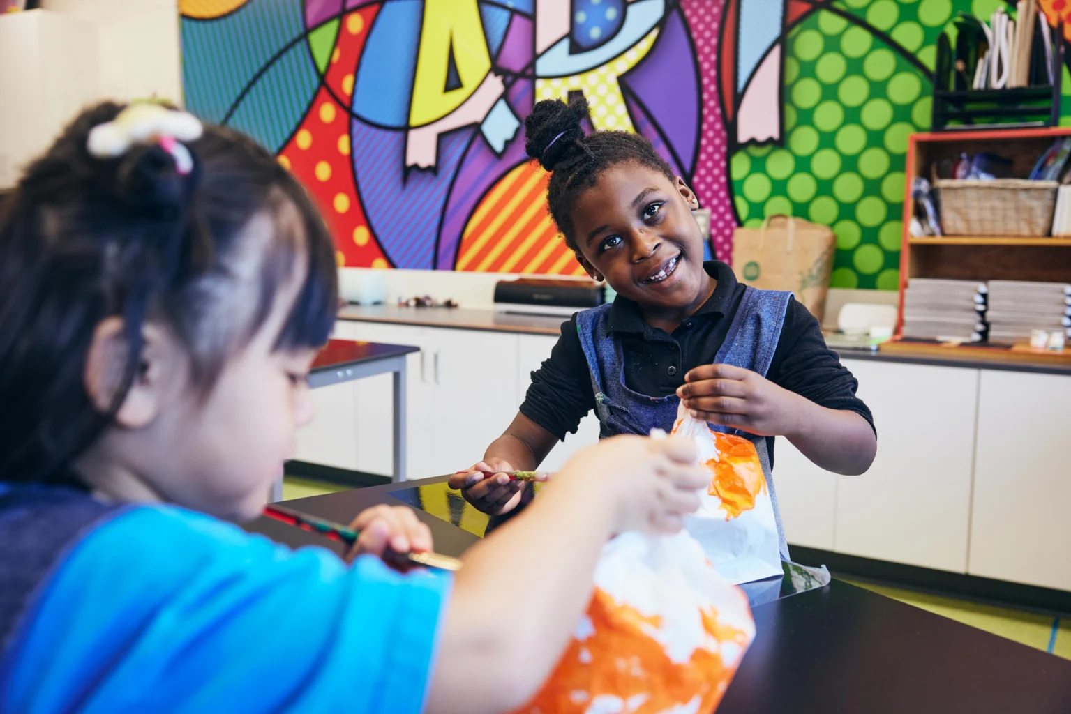 Two children work on colorful paper crafts at a table in an art classroom, with bright paintings and supplies visible in the background.