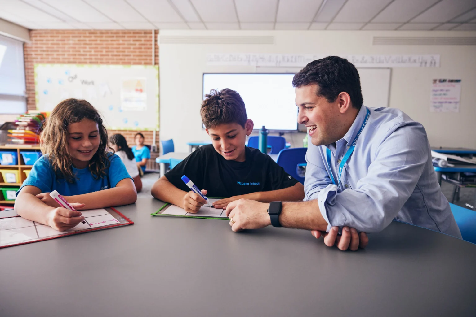 A teacher sits at a table with two students, helping them with their work in a brightly lit classroom.