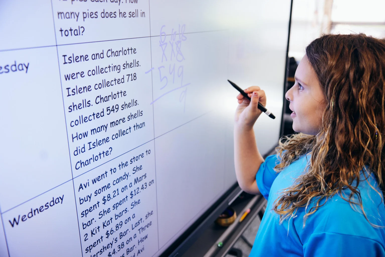 A girl writes math calculations with a stylus on an interactive whiteboard displaying a word problem about shell collection.