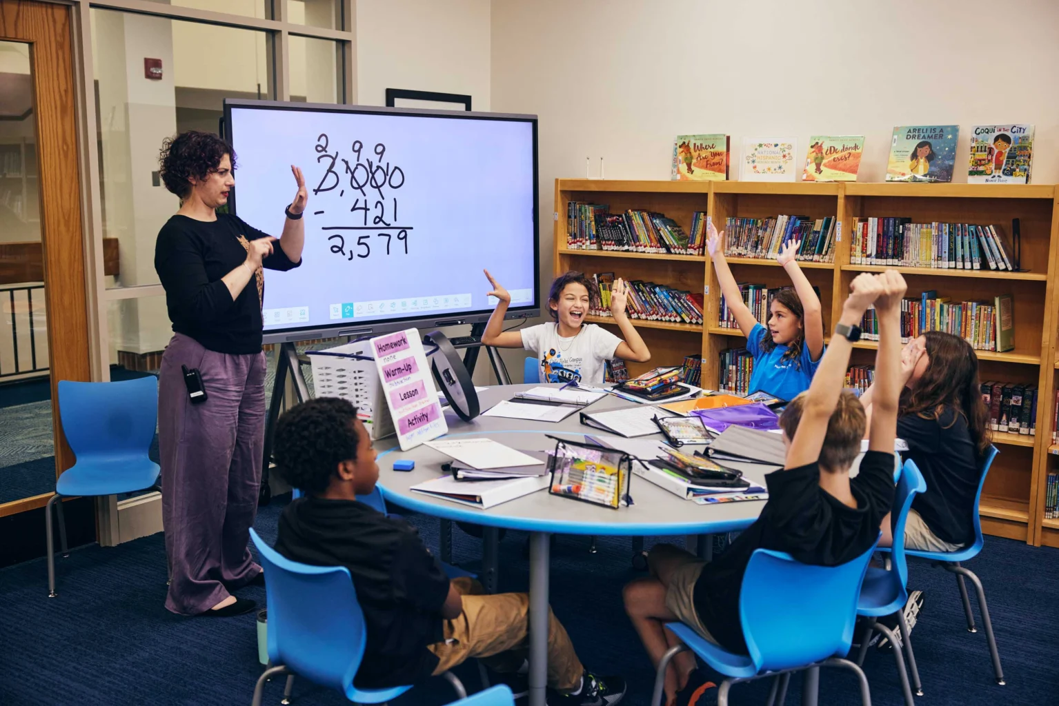 A teacher stands by a screen displaying math problems while five students sitting at a table raise their hands in a classroom with bookshelves in the background.