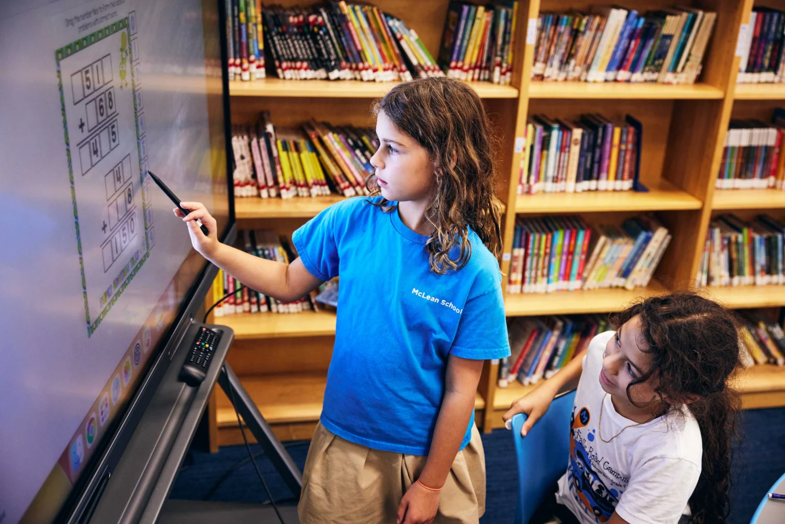Two children work on math problems displayed on a large touchscreen in a library, with bookshelves filled with books in the background.