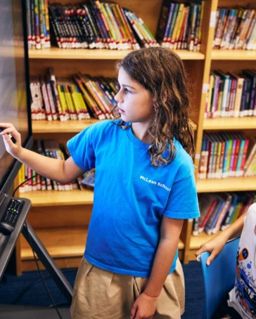 Two children work on math problems displayed on a large touchscreen in a library, with bookshelves filled with books in the background.
