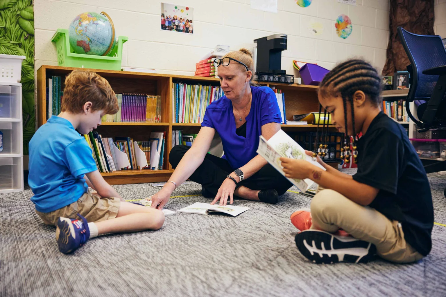 A teacher sits on the floor with two young students in a classroom, helping them with reading activities and books.
